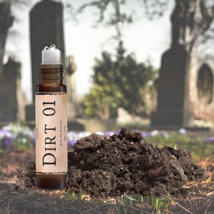 May include: A brown glass roll-on perfume bottle with a white cap. The label is brown and white and says "DIRT 01" and "WITCHERY" in a vintage font. The bottle is sitting on a pile of dirt in front of a blurry background of a graveyard.