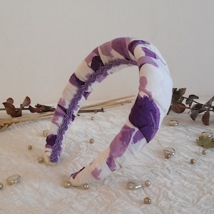 May include: A purple and white patterned headband with a ruffled trim. The headband is made of fabric and is designed to be worn around the head.