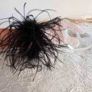 Puede incluir: Un brazalete de plumas negras combinado con un guante de red blanco. El brazalete presenta un denso grupo de largas plumas negras. El guante está hecho de una tela delicada de tejido abierto. Los artículos se exhiben sobre una superficie blanca texturizada.