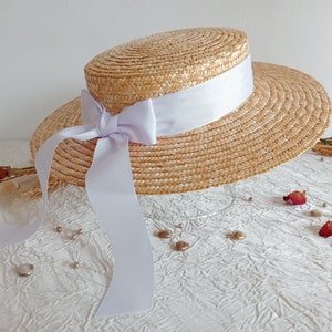 Peut inclure: Un chapeau de paille à large bord avec un ruban de satin blanc noué en un nœud. Le chapeau est posé sur une surface blanche avec de petits objets décoratifs.