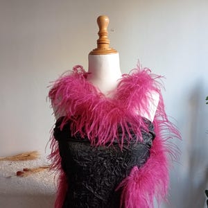 Op de afbeelding: Een felroze verenboa over een zwarte jurk op een mannequin. De boa is pluizig en levendig, wat contrasteert met de donkere textuur van de jurk. De mannequin staat tegen een neutrale achtergrond, waardoor de kleur en textuur van de boa worden benadrukt.