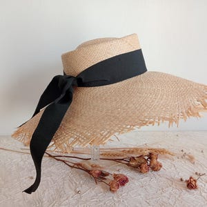Op de afbeelding: Een natuurlijke strohoed met een brede rand en een zwart lint dat in een strik is geknoopt. De hoed heeft een gerafelde rand en een cilindrische kroon. Een modieus accessoire voor bescherming tegen de zon.