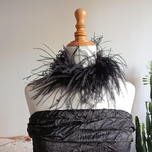 May include: A black feather boa collar on a mannequin. The collar is made of soft, dark feathers and sits around the mannequin's neck. The mannequin is wearing a black, textured garment. A wooden stand supports the mannequin.
