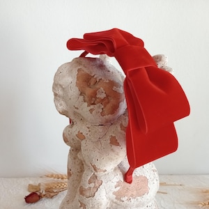 Puede incluir: Una diadema con lazo de terciopelo rojo brillante. El lazo es grande y llamativo, con un aspecto suave y texturizado. Esta diadema es un accesorio de moda, ideal para añadir un toque de color a cualquier atuendo.