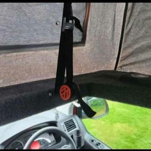 May include: Interior of a toy car, showing a black seatbelt with a red and black buckle. The dashboard and steering wheel are visible, with a green lawn visible through the windshield and side mirror.