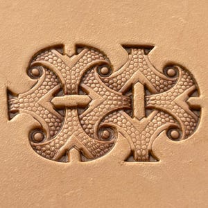 May include: A tan leather surface featuring a deeply embossed, symmetrical pattern. The design consists of two mirrored, ornate shapes with textured, pebbled centers, smooth raised borders, and small circular accents at each of the four corners.