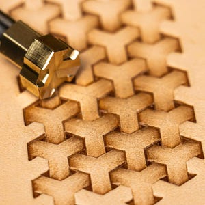 May include: A close-up shot of a leatherworking tool with a brass head, impressing a geometric pattern onto tan leather. The tool is used to create a repeating Y-shaped design. The image shows the detail of the tooling process.