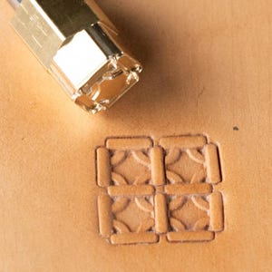May include: A close-up of a leather stamping tool with a geometric design. The tool is made of gold-coloured metal and is pressed into a piece of tan leather, creating an embossed pattern. The stamped design is a repeating square pattern.