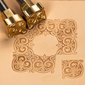 Puede incluir: Primer plano de dos sellos de cuero de metal dorado con un diseño de búho, junto a un patrón en relieve detallado sobre cuero marrón claro. El diseño en relieve presenta un marco cuadrado con detalles florales y de volutas.