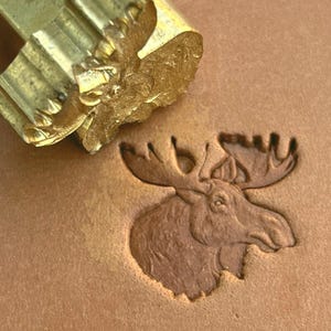 May include: A gold-toned metal leather stamping tool featuring a detailed moose head design. The tool is positioned above a tan leather surface that displays a clear, recessed impression of the same moose head, showing the antlers and snout.