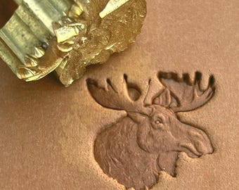 May include: A gold-toned metal leather stamping tool featuring a detailed moose head design. The tool is positioned above a tan leather surface that displays a clear, recessed impression of the same moose head, showing the antlers and snout.
