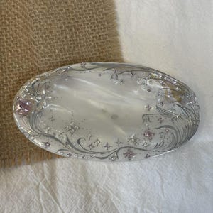 May include: An oval-shaped tray with a pearlescent white finish, decorated with silver filigree patterns of swirling vines and small flowers. The surface is accented with embedded clear and pink crystals arranged in star and floral motifs.