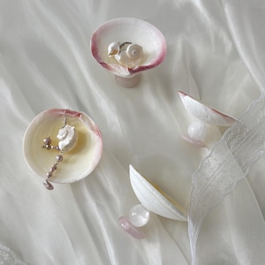 May include: Four small decorative dishes crafted from white and pink-rimmed seashells, each supported by a clear or pale pink stone base. One dish holds a silver ring, another contains a dangling earring with small mauve pearls.