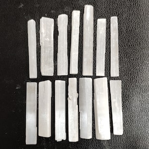 Puede incluir: Una colección de 16 varillas de selenita blanca, dispuestas en dos filas. Las varillas son lisas y tienen una textura natural y cristalina.