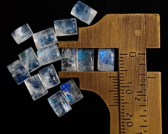 May include: A collection of iridescent blue moonstone rectangular cabochons, measured with a ruler in inches. The stones are translucent and have a milky white color with a blue sheen.