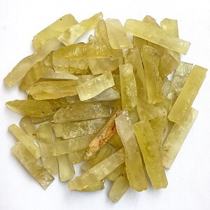 Puede incluir: Una colecci&oacute;n de puntas de cristal natural en bruto de color amarillo. Las puntas son largas y delgadas, con una apariencia transl&uacute;cida. Los cristales est&aacute;n dispuestos en un mont&oacute;n suelto sobre un fondo blanco.