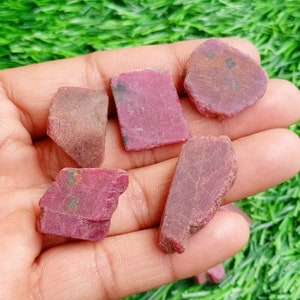 Può includere: Cinque pezzi di pietra preziosa di rubino rosso grezzi in varie forme e dimensioni. Le pietre sono grezze e non levigate, con un aspetto naturale e terroso.