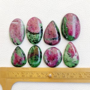 May include: Eight green and pink gemstone cabochons, some oval shaped and some teardrop shaped, arranged on a white surface with a gold ruler for scale. The ruler is marked in inches.