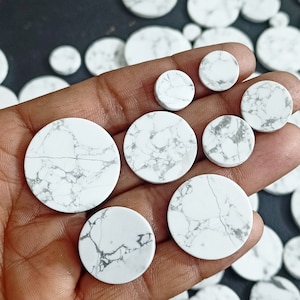 Può includere: Una collezione di cabochon rotondi in howlite bianca di diverse dimensioni. I cabochon hanno un motivo marmoreo con venature grigie.