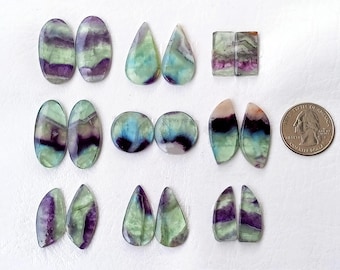 Puede incluir: Surtido de 12 cabujones de fluorita verde, morado y azul en varias formas, incluyendo ovalado, lágrima y rectangular. Los cabujones están dispuestos en filas sobre una superficie blanca. Se incluye una moneda de veinticinco centavos de dólar estadounidense para la escala.
