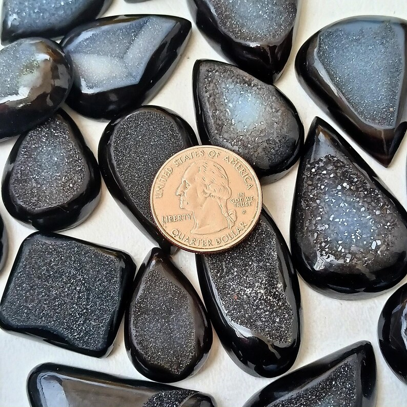 Op de afbeelding: Een verzameling zwarte en grijze agaat cabochons met een glinsterende, iriserende afwerking. De cabochons zijn in verschillende vormen en maten, waaronder druppelvormig, ovaal en rechthoekig. Een Nederlandse euro is opgenomen voor de schaal.