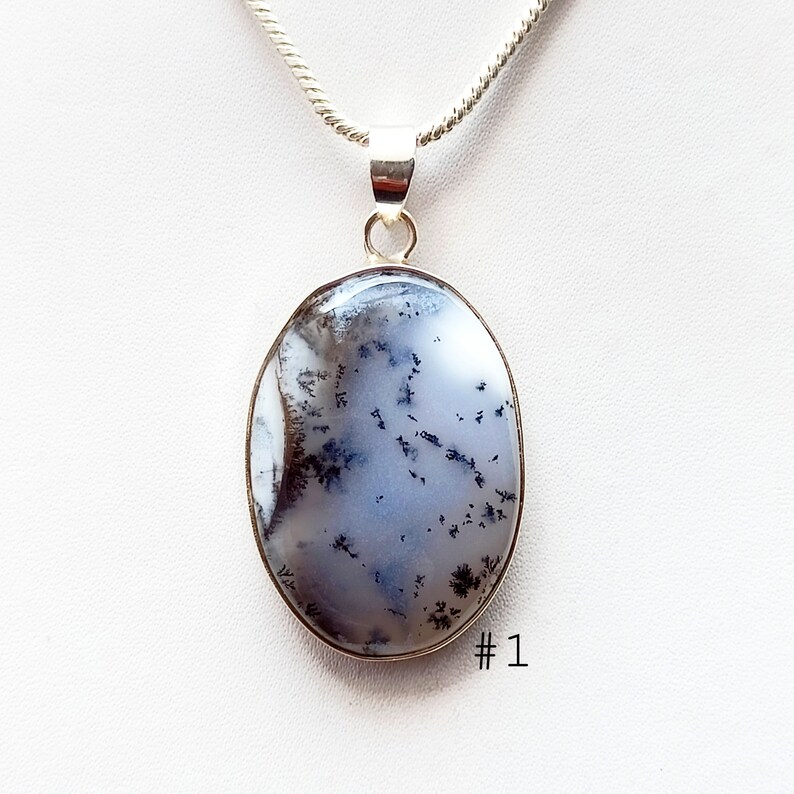 May include: Oval pendant with a silver-colored metal frame and a dendritic agate stone. The stone features a white and light blue background with black and brown tree-like patterns. The pendant hangs from a silver chain. The number "1" is printed in the lower right corner.