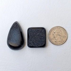 Op de afbeelding: Twee zwarte edelsteen cabochons, een traanvormig en een vierkant, naast een Amerikaanse kwart dollar munt voor groottevergelijking.