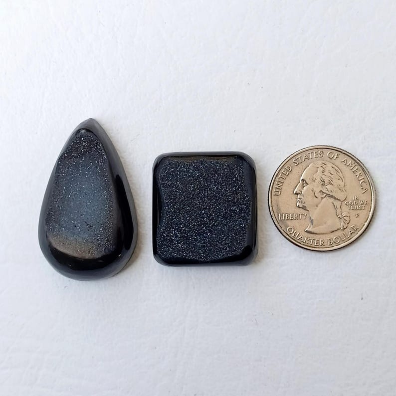 Op de afbeelding: Twee zwarte edelsteen cabochons, een traanvormig en een vierkant, naast een Amerikaanse kwart dollar munt voor groottevergelijking.