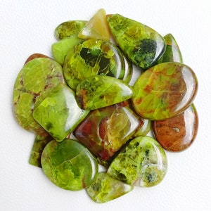 May include: A collection of polished gemstones in shades of green, yellow, and brown. The stones vary in shape, including oval, teardrop, and rectangular. Some stones have a mottled appearance with darker green and brown inclusions. The gemstones are arranged on a white surface.