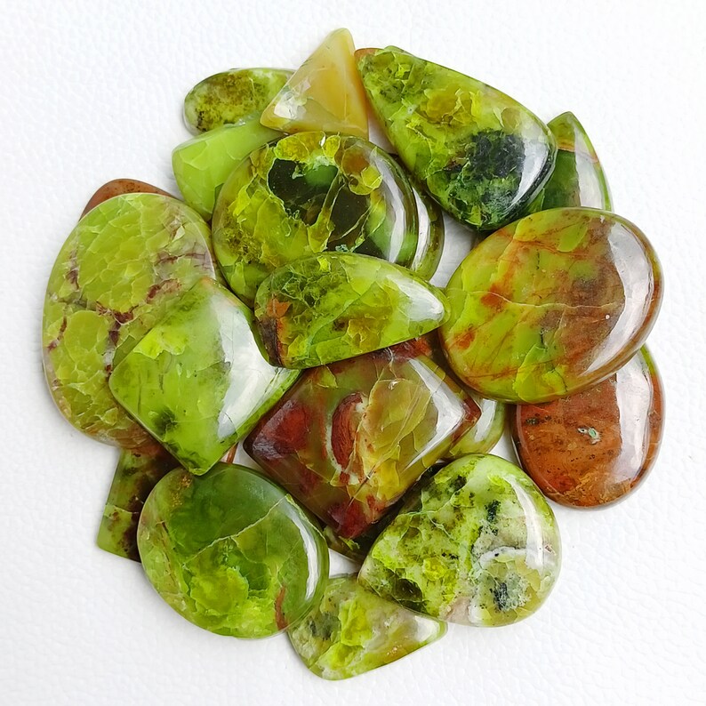 May include: A collection of polished gemstones in shades of green, yellow, and brown. The stones vary in shape, including oval, teardrop, and rectangular. Some stones have a mottled appearance with darker green and brown inclusions. The gemstones are arranged on a white surface.
