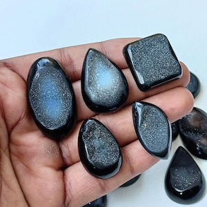 Op de afbeelding: Een verzameling van zes zwarte en witte agaat-cabochons. De cabochons zijn in verschillende vormen, waaronder druppel, ovaal en vierkant. De cabochons hebben een glinsterende, iriserende afwerking.