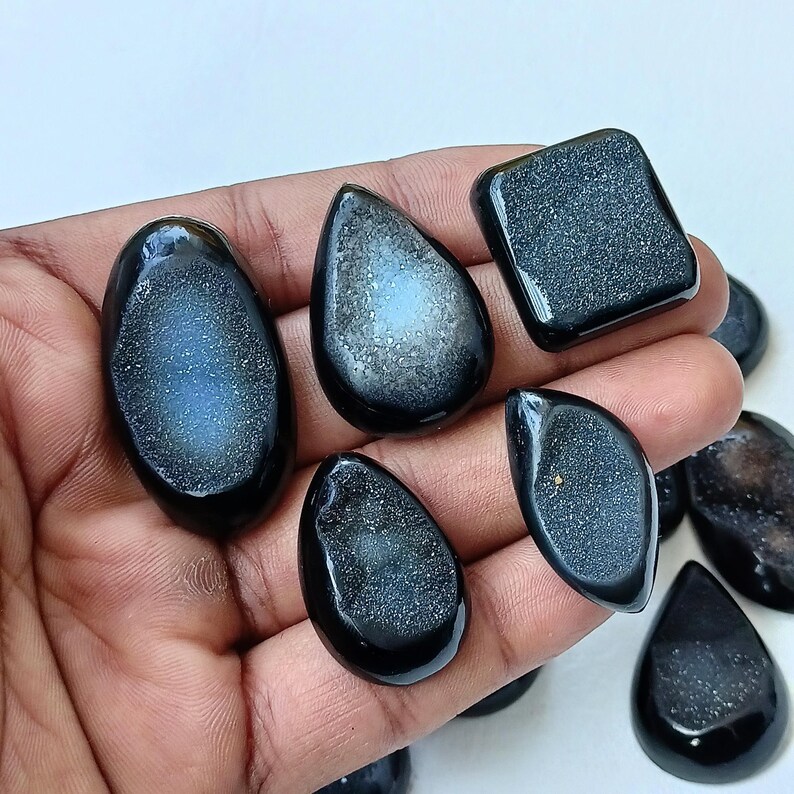 Op de afbeelding: Een verzameling van zes zwarte en witte agaat-cabochons. De cabochons zijn in verschillende vormen, waaronder druppel, ovaal en vierkant. De cabochons hebben een glinsterende, iriserende afwerking.