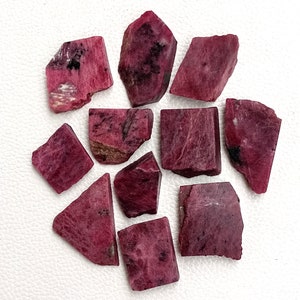 May include: A collection of ten rough, pink ruby gemstones. The stones are various shapes and sizes, with some having a more polished look than others.