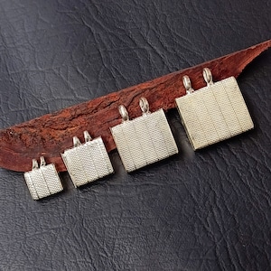 May include: Five silver-toned metal pendant charms of varying sizes, each with a square shape and a textured surface. The charms are arranged in a row on a brown wooden surface.