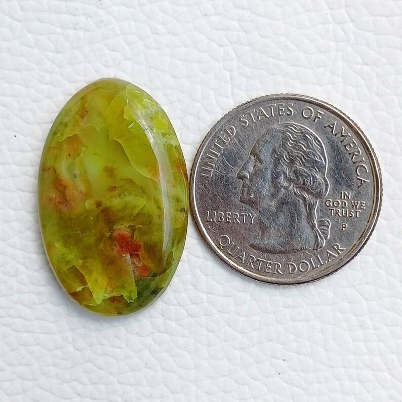 May include: An oval-shaped gemstone with a mix of green, yellow, and red hues. The gemstone is next to a US quarter coin for size comparison. The stone has a polished, glossy surface.