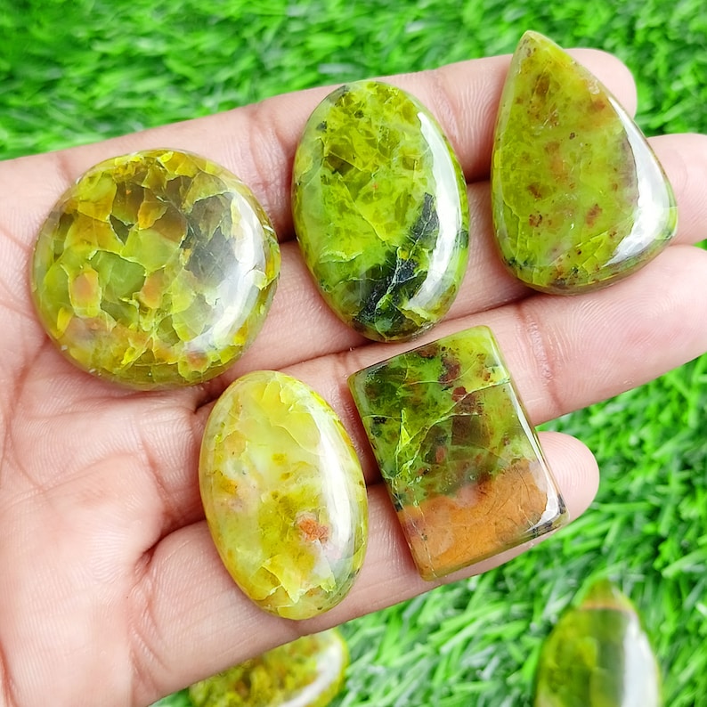 May include: A collection of polished gemstones in various shapes, including oval, round, and teardrop. The stones display a vibrant green color with brown and yellow inclusions, creating a unique, natural pattern. The gemstones are displayed on a green surface.