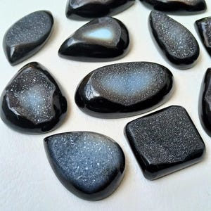 Op de afbeelding: Een verzameling zwarte agaat cabochons met een wit druzy interieur. De cabochons zijn in verschillende vormen en maten en hebben een gepolijste afwerking.
