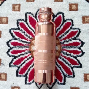 May include: A copper water bottle with a hammered finish and a silver lid. The bottle has a traditional design with horizontal bands.