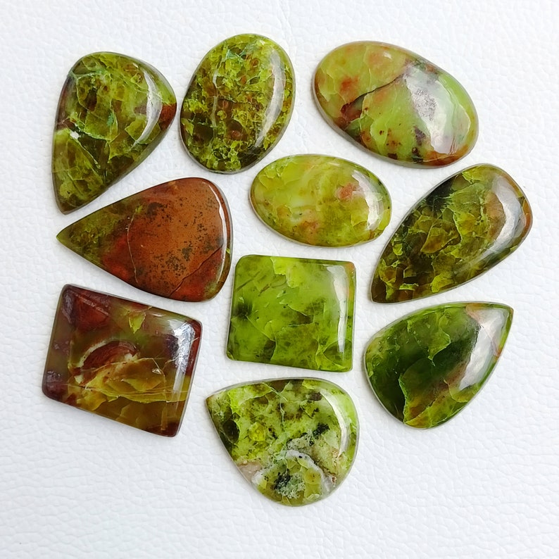 May include: A collection of polished gemstones in various shapes, including oval, teardrop, and rectangular. The stones display a mix of green and brown hues, with some featuring a mottled or veined appearance. The gemstones are arranged on a white surface.