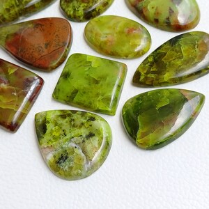 May include: A collection of polished gemstones in various shapes, including oval, square, and teardrop. The stones display a mix of green and brown hues, with some featuring intricate patterns and textures. The gemstones are arranged on a white surface.