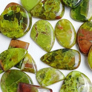 May include: A collection of polished gemstones in various shapes, including ovals, rectangles, and freeform designs. The stones display a range of greens, from light lime to deep olive, with some showing reddish-brown inclusions. The surfaces are smooth and glossy.