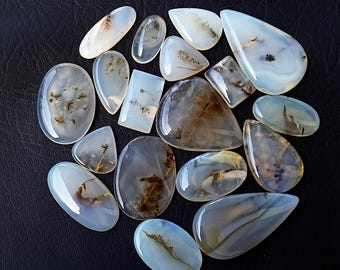 May include: A collection of polished dendritic agate gemstones in various shapes, including oval, teardrop, and rectangular. The stones feature translucent white and pale blue hues with brown and black inclusions, set against a dark background.