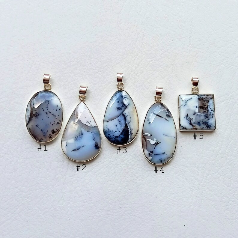 May include: Five dendritic opal pendants with silver-colored bails. The pendants vary in shape, including oval, teardrop, and square. Each pendant features a unique pattern of white, gray, and black inclusions within the opal.