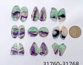 Puede incluir: Una colección de 12 cabujones de fluorita de color verde, morado y blanco. Los cabujones son de varias formas y tamaños, incluyendo forma de lágrima, ovalada y rectangular. Los cabujones están dispuestos en pares sobre una superficie blanca.