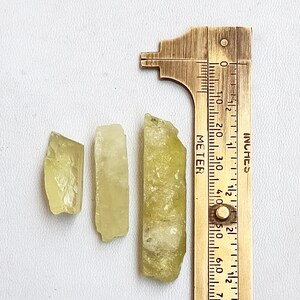 Puede incluir: Tres cristales de color amarillo verdoso de diferentes tama&ntilde;os, medidos con una regla de lat&oacute;n marcada en pulgadas. El cristal m&aacute;s grande mide aproximadamente 1 1/2 pulgadas de largo.