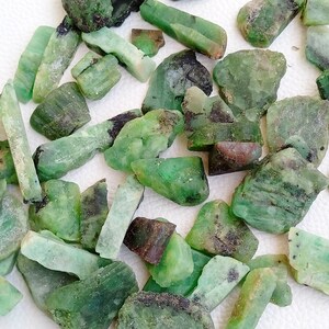 Può includere: Una collezione di pietre preziose di smeraldo verde grezzo. Le pietre sono di varie dimensioni e forme, alcune hanno una tonalità verde più scura.