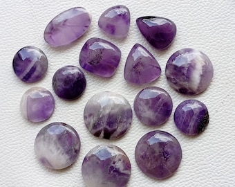 Könnte beinhalten: Eine Sammlung von 15 Amethyst-Cabochons in verschiedenen Formen und Größen. Die Cabochons sind eine Mischung aus Lila und Weiß, wobei einige ein ausgeprägteres weißes Muster aufweisen.