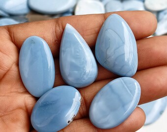 May include: Five blue opal cabochons in various shapes, including oval and teardrop, with a smooth, polished finish. The stones are held in a hand, showcasing their light blue color and translucent quality.