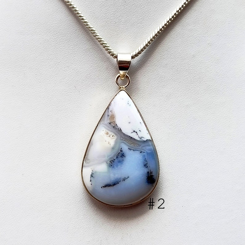 May include: A teardrop-shaped pendant with a silver chain. The pendant features a large, polished stone with a white, gray, and blue pattern. The stone is set in a silver bezel. The number "2" is visible in the lower right corner.