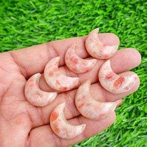 May include: Seven sunstone crescent moon shaped crystals in various shades of orange and white. The crystals are arranged in a hand.